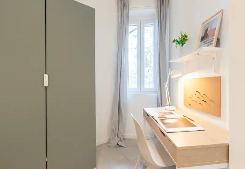 Coin de chambre compact avec armoire et bureau en bois clair sous la fenêtre ; style minimaliste et lumineux, avec panneau en liège, lampe de bureau et rideaux doux.