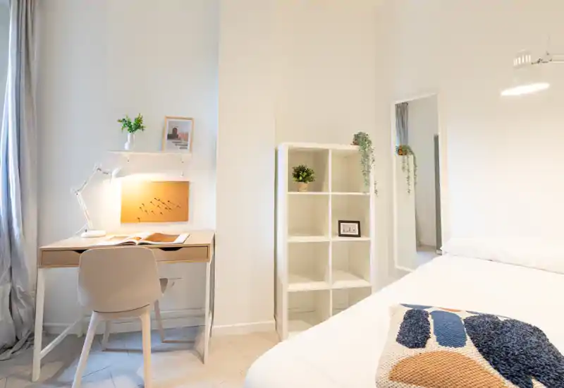 Rincón de dormitorio moderno y luminoso con cama cómoda, cojín decorativo y un escritorio ordenado con lámpara — estilo minimalista y relajante.