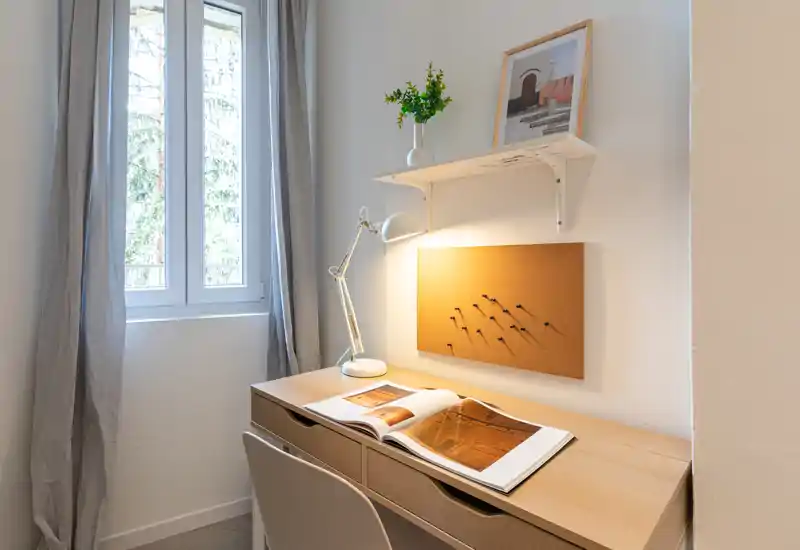 Coin bureau lumineux et minimaliste avec un meuble en bois, une lampe articulée, un panneau de liège et un livre ouvert près de la fenêtre — parfait pour un petit espace bureau ou coin étude.