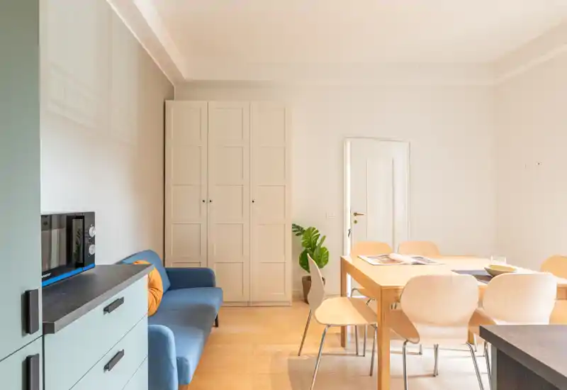 Espace de vie lumineux et moderne avec une table à manger en bois, six chaises claires et un canapé bleu compact. Sol en bois chaud, murs neutres et une plante en pot pour une atmosphère accueillante.