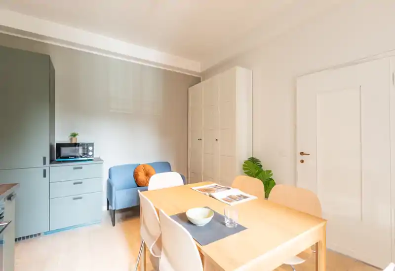 Coin salon/salle à manger lumineux et bien agencé avec une table en bois, quatre chaises, un petit canapé bleu et des rangements intégrés — idéal pour un appartement compact.