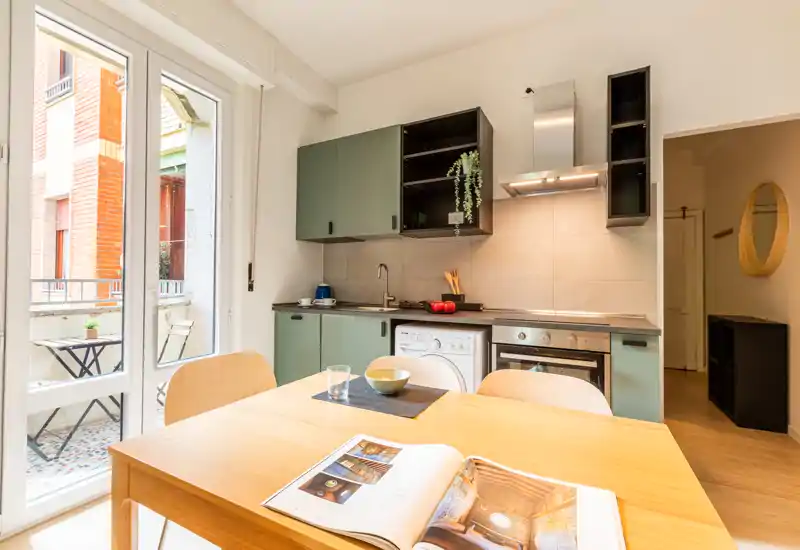 Cuisine compacte et lumineuse avec placards verts, four encastré et lave-linge, table en bois près de la porte du balcon — idéale pour un séjour en ville.
