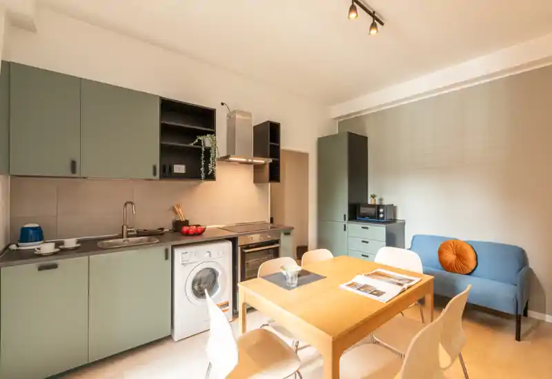 Cuisine compacte et moderne avec appareils intégrés, table en bois et petit coin salon. Les façades vertes et l'éclairage chaleureux créent un espace accueillant et fonctionnel, parfait pour des séjours courts.