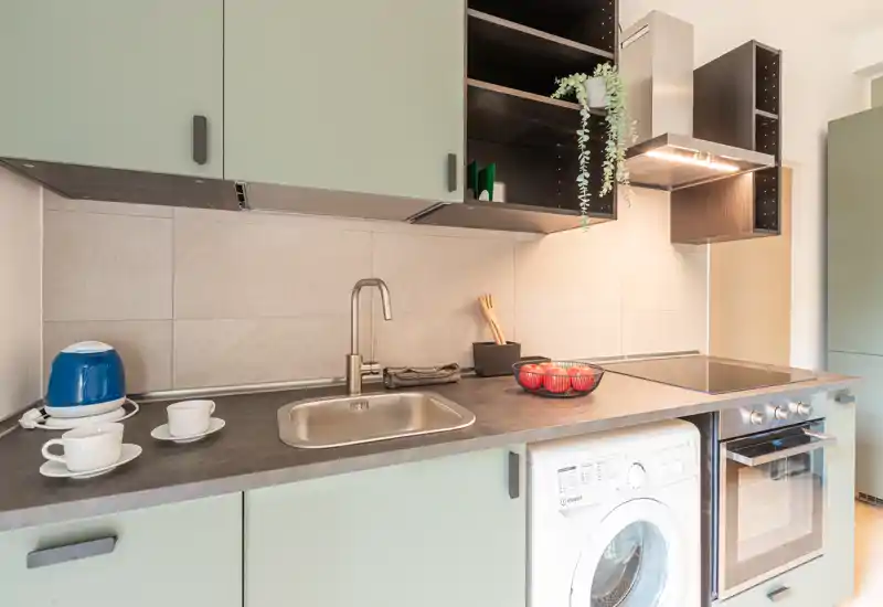 Cuisine compacte et moderne avec placards vert menthe, four intégré et lave-linge, évier en acier inoxydable et plaque vitrocéramique. Espace lumineux et bien mis en scène avec des touches décoratives (bol de fruits, plante).