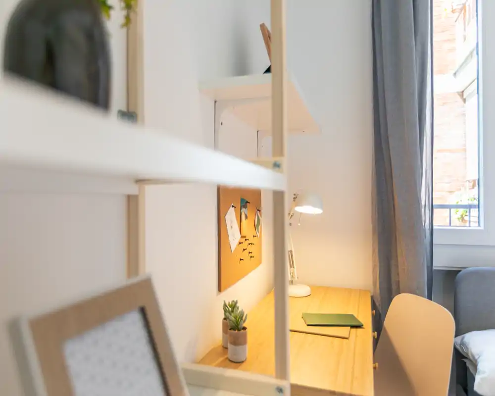Angolo di lavoro accogliente e ben illuminato con scrivania in legno, lampada da tavolo, bacheca e una piccola pianta vicino alla finestra — ideale per studio o smart working.