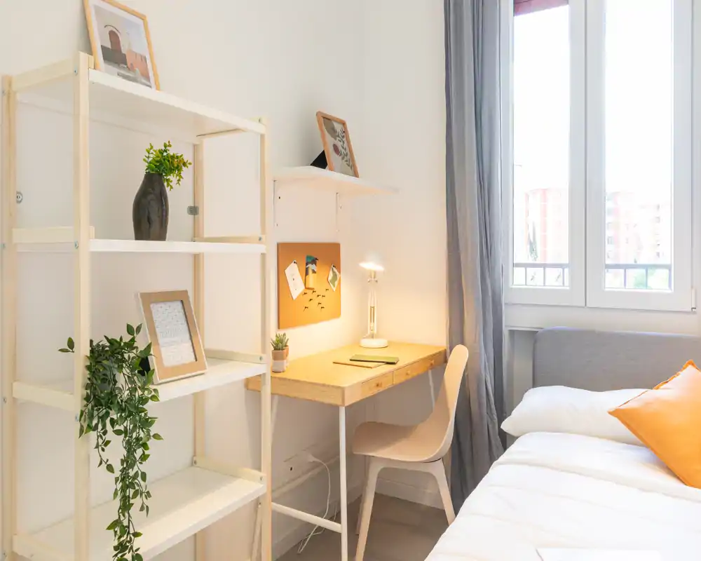 Angolo camera luminoso e accogliente con letto comodo, cuscini e una piccola scrivania in legno vicino alla finestra — perfetto per riposare e lavorare.