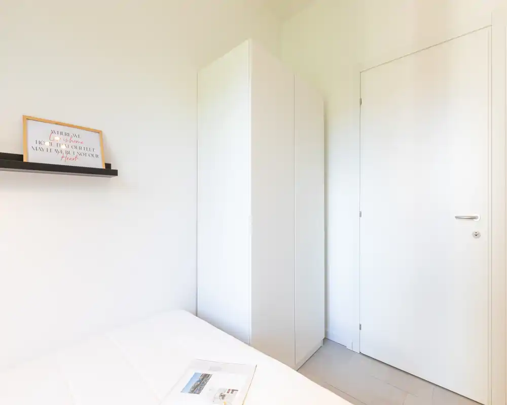 Angolo di camera da letto luminoso e compatto con un armadio bianco e letto ordinato. Arredamento minimal con una mensola a parete con quadro e luce naturale morbida.