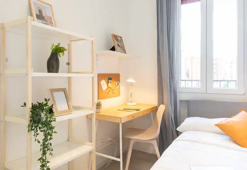 Angolo camera luminoso e accogliente con letto comodo, cuscini e una piccola scrivania in legno vicino alla finestra — perfetto per riposare e lavorare.