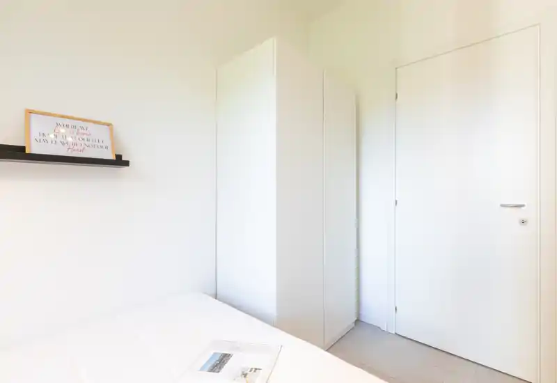 Angolo di camera da letto luminoso e compatto con un armadio bianco e letto ordinato. Arredamento minimal con una mensola a parete con quadro e luce naturale morbida.