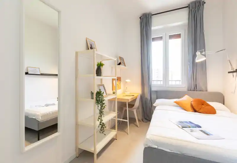 Camera da letto luminosa e ben arredata con comodo letto matrimoniale, scrivania vicino alla finestra e cuscini decorativi — perfetta per un soggiorno accogliente.