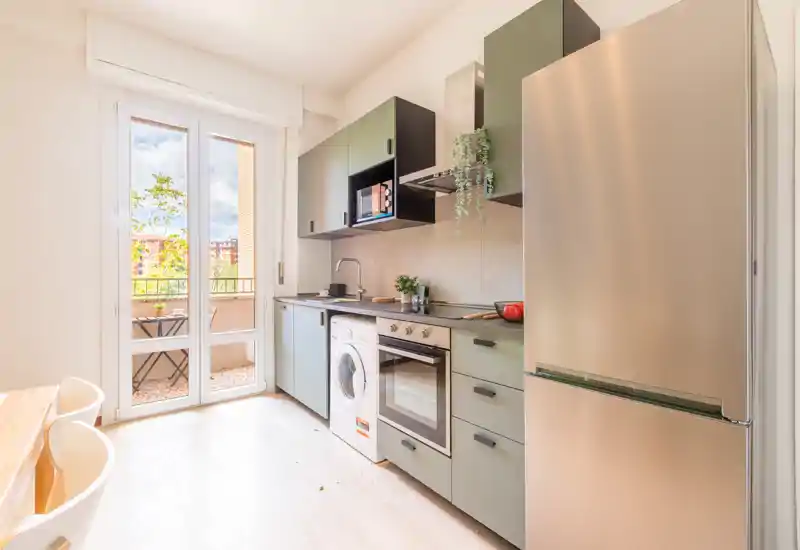 Cuisine moderne et lumineuse avec appareils intégrés, grand réfrigérateur, four et lave-linge, et une porte donnant sur le balcon apportant beaucoup de lumière naturelle.