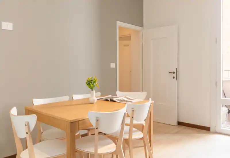 Coin repas lumineux et minimaliste avec une table en bois pour six et des chaises blanches modernes ; la lumière naturelle et un petit vase ajoutent une touche accueillante.
