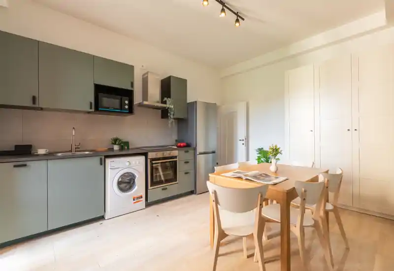 Cuisine moderne et lumineuse avec électroménagers intégrés et une table en bois pour quatre personnes ; placards verts, lave-linge, four et réfrigérateur forment un espace de cuisson et de repas fonctionnel et stylé.