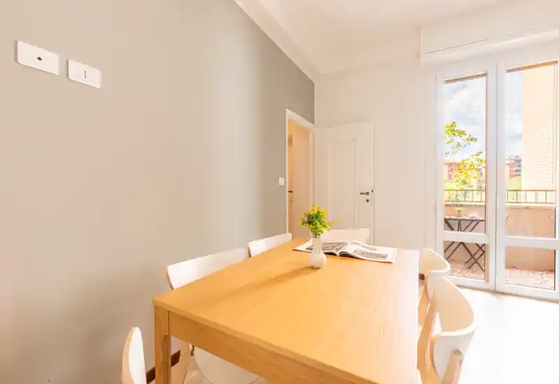 Coin repas lumineux et moderne avec table en bois, chaises blanches et porte-fenêtre donnant sur le balcon ; décoration simple avec un petit vase et un magazine ouvert.