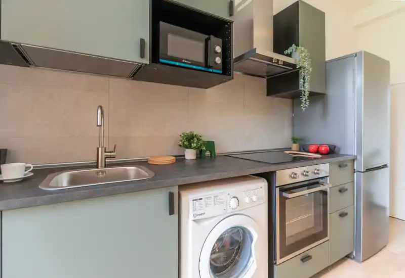 Cuisine compacte et moderne avec électroménagers intégrés : four, plaque à induction, micro-ondes, réfrigérateur et lave-linge sous le plan de travail. Un dosseret neutre, des meubles aux teintes douces et de petites plantes créent un espace propre et chaleureux.