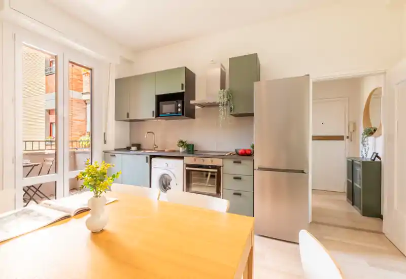 Cuisine ouverte moderne et lumineuse avec meubles verts, électroménagers inox et table en bois. Une grande baie donne sur un balcon, apportant lumière et une atmosphère agréable pour les repas.