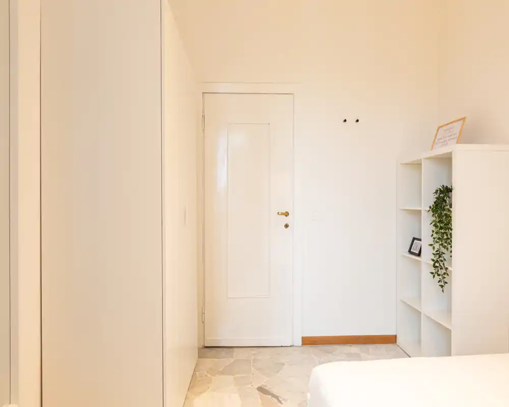 Angolo di camera da letto luminoso e minimal: porta bianca, armadio e parziale vista del letto; tonalità neutre e scaffalatura semplice con una piccola pianta.