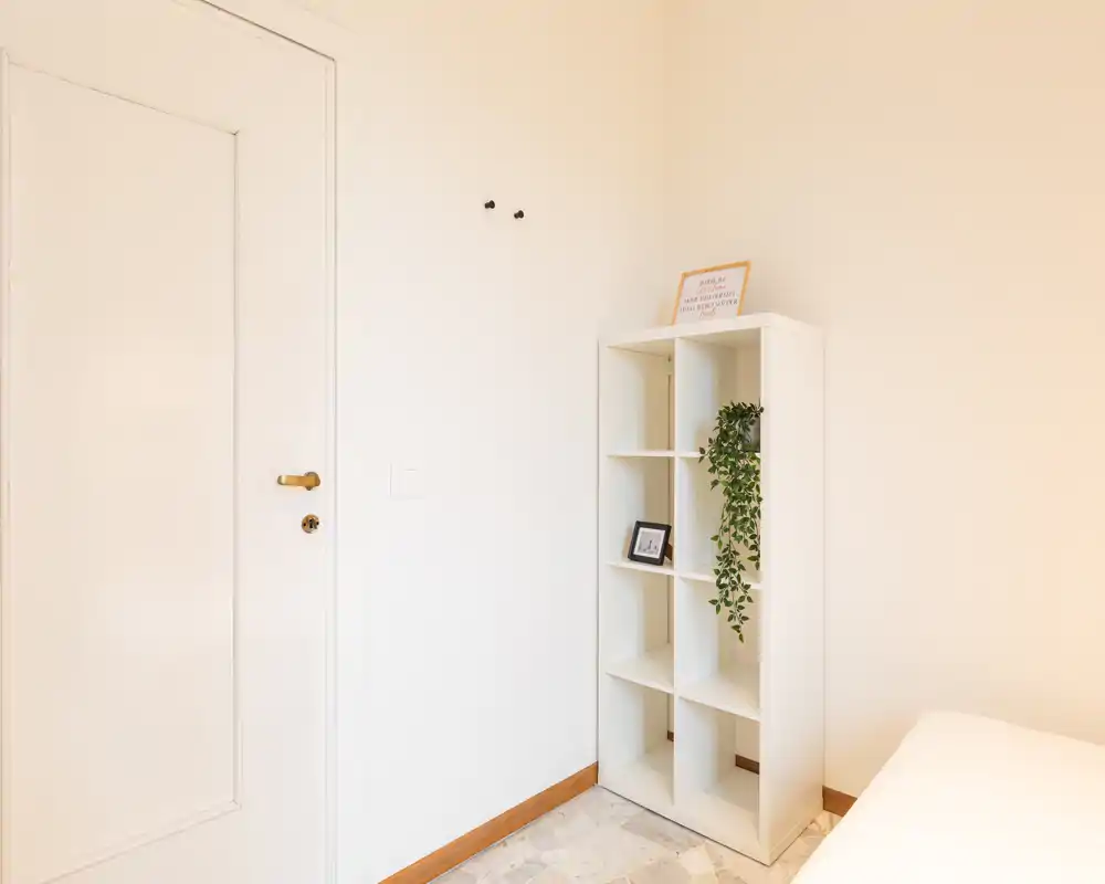 Angolo di camera da letto luminoso e minimal con una libreria bianca, una piccola pianta decorativa e il letto parzialmente visibile sul bordo dell'inquadratura. Lo spazio risulta pulito, ben illuminato e arredato con semplicità — ideale per mettere in risalto luce e spazio di stoccaggio.