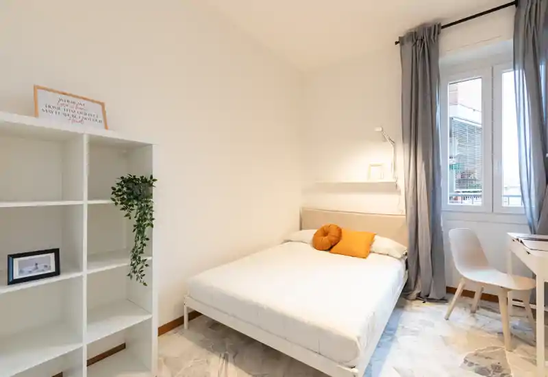 Camera da letto luminosa e moderna con un letto matrimoniale ben rifatto, toni neutri e una finestra che lascia entrare luce naturale. Mensole semplici e una piccola scrivania rendono lo spazio funzionale e minimal, ideale per soggiorni brevi.