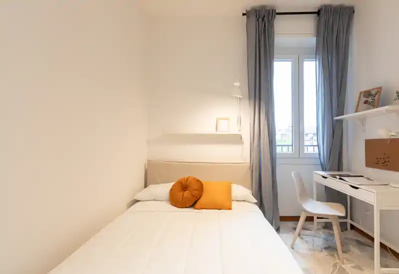 Camera da letto luminosa e compatta con letto ben rifatto, cuscini dagli accenti caldi e una piccola scrivania vicino alla finestra — pulita, luminosa e ben arredata per soggiorni brevi.