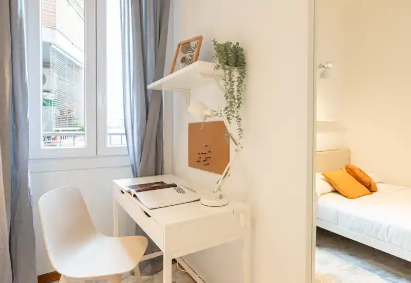 Angolo camera luminoso e compatto con una scrivania funzionante vicino alla finestra e un letto parzialmente visibile con cuscini colorati.
