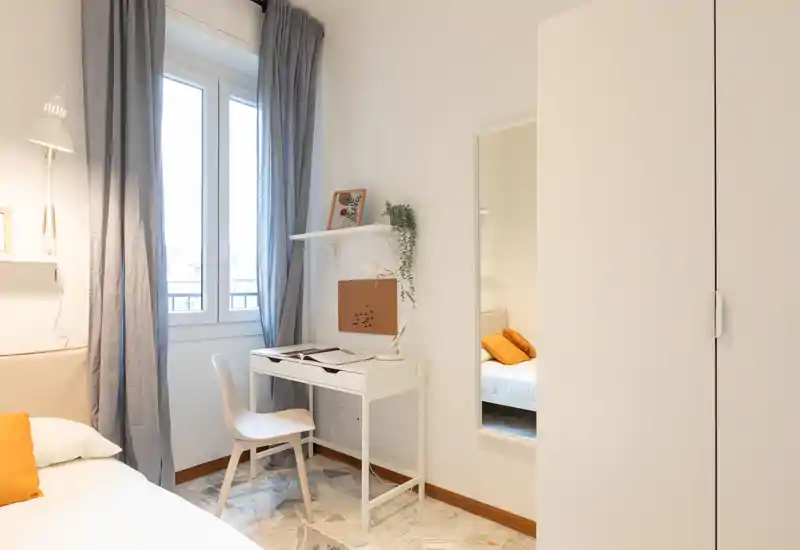 Angolo camera luminoso e curato con letto singolo, lampada, scrivania e armadio; la luce naturale dalla finestra e gli accenti caldi rendono l’ambiente accogliente.