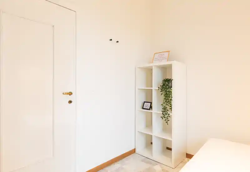 Rincón de dormitorio luminoso y minimalista con una estantería blanca, una pequeña planta decorativa y la cama parcialmente visible en el borde del encuadre. El espacio se ve limpio, bien iluminado y discretamente amueblado — ideal para destacar la luz y el almacenaje.