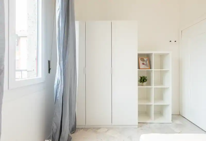 Bright, minimal bedroom corner with a white wardrobe and open shelving, natural light from a nearby window and light marble floor.