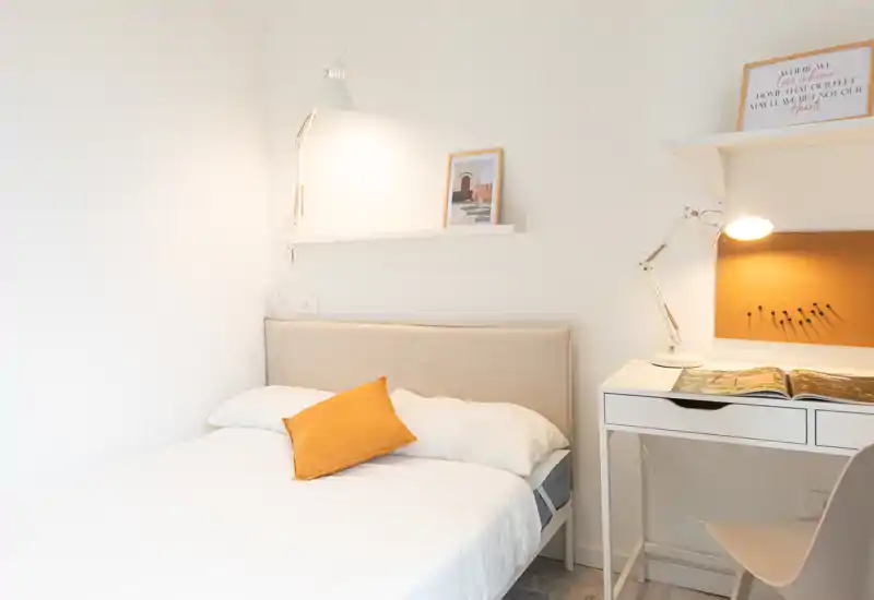 Bright, minimal bedroom with a neatly made single bed, warm accent pillow, and a compact workspace with task lighting — ideal for a comfortable solo stay.