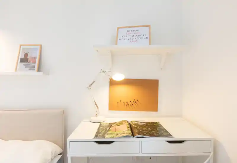 Minimal, bright bedroom corner with a bedside desk and lamp; a neatly made bed is partially visible on the left, and decorative shelves and a cork board create a calm, airy atmosphere.