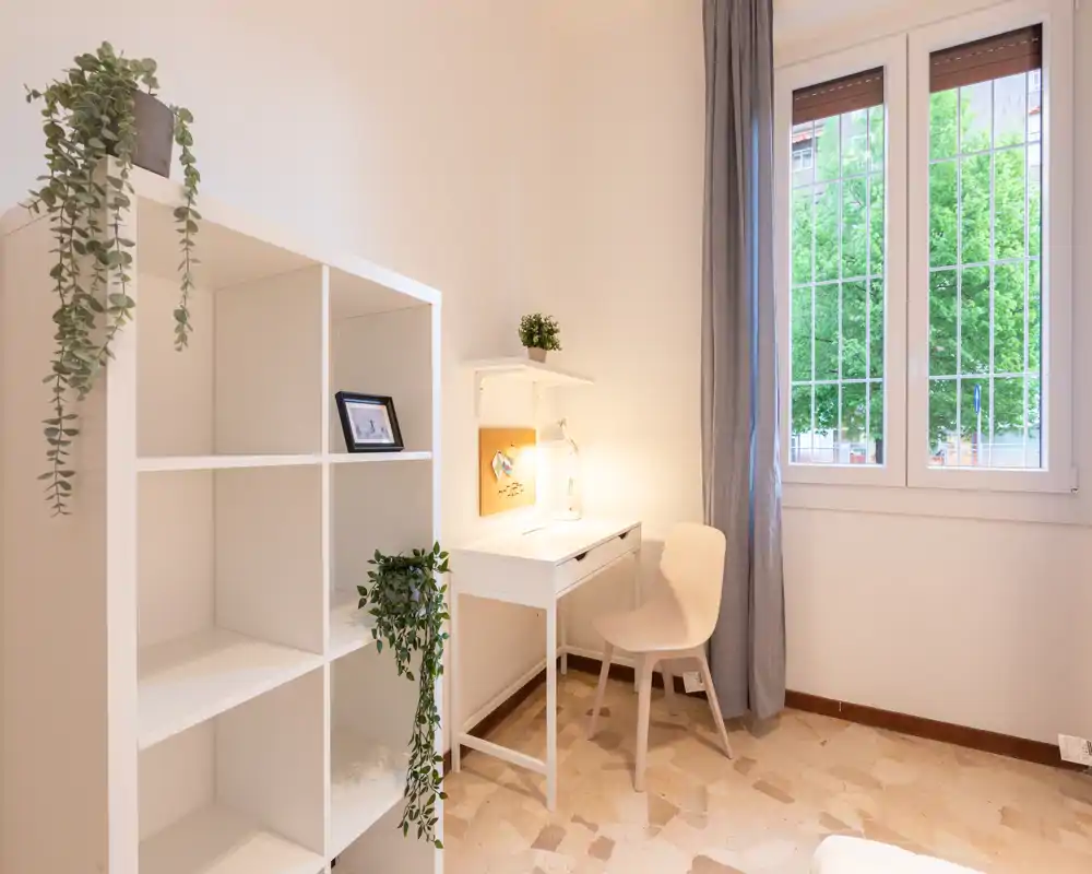 Spazio giorno luminoso e ordinato con una piccola scrivania vicino alla finestra, scaffalatura minimal e piante decorative — ideale per studiare o rilassarsi con luce naturale.