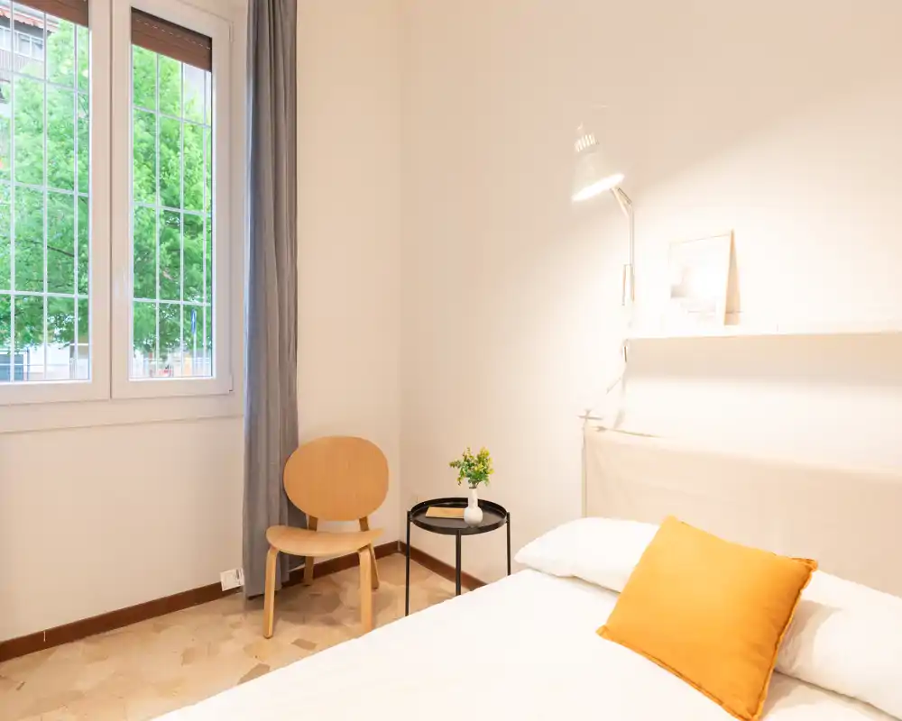 Camera da letto luminosa e minimalista con letto ordinato, cuscino dai toni caldi e lampada da comodino. Una sedia semplice e un tavolino con una pianta vicino alla finestra creano un'atmosfera tranquilla e ariosa, perfetta per annunci.