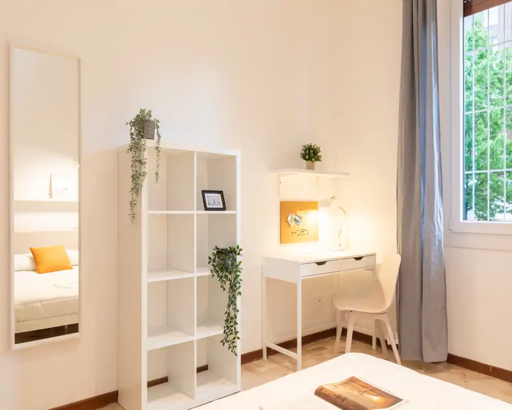 Camera da letto luminosa e minimale con letto ben visibile, tessili accoglienti, una scrivania con mensole e luce naturale da una finestra ampia.