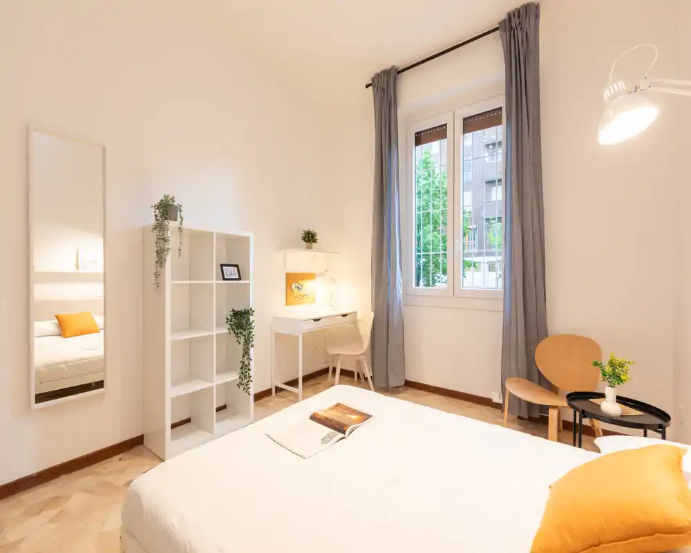 Camera da letto luminosa e di stile con letto matrimoniale ben rifatto, biancheria morbida e cuscini colorati. La stanza dispone di una scrivania vicino alla finestra, scaffalatura e un angolo salotto accogliente.