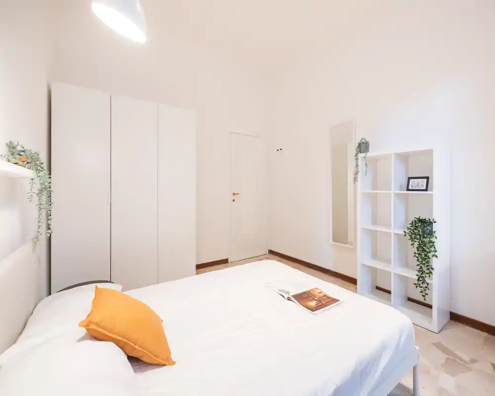 Camera da letto luminosa e minimal con letto ampio con lenzuola bianche, cuscino accent arancione, armadio e scaffale bianchi e piccole piante che aggiungono un tocco fresco.
