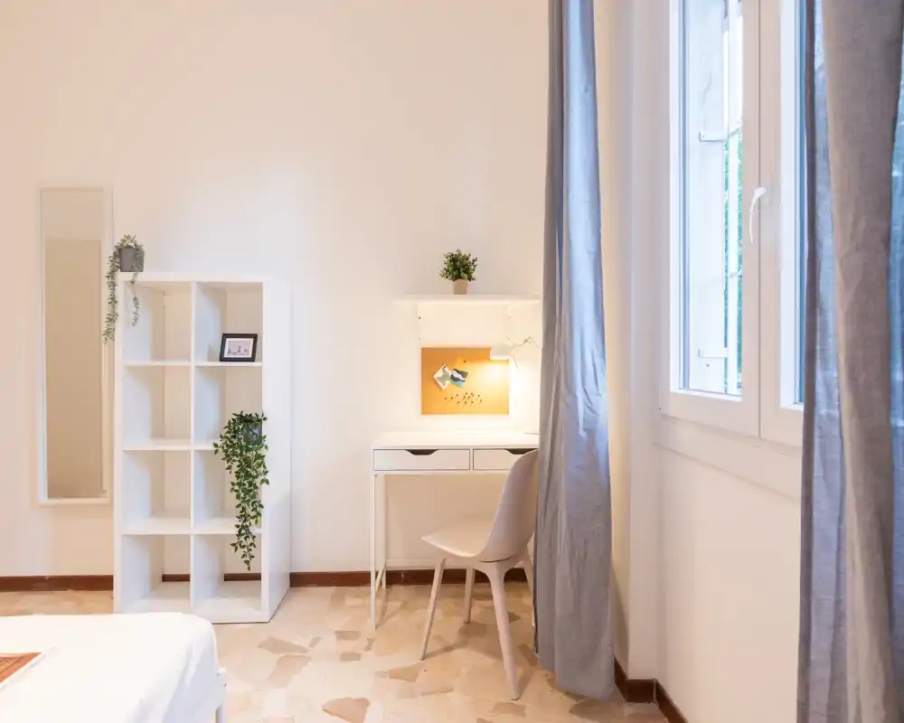 Angolo di camera minimal e luminoso con una piccola porzione del letto visibile, scrivania bianca, scaffale, piante e una grande finestra che illumina la stanza.