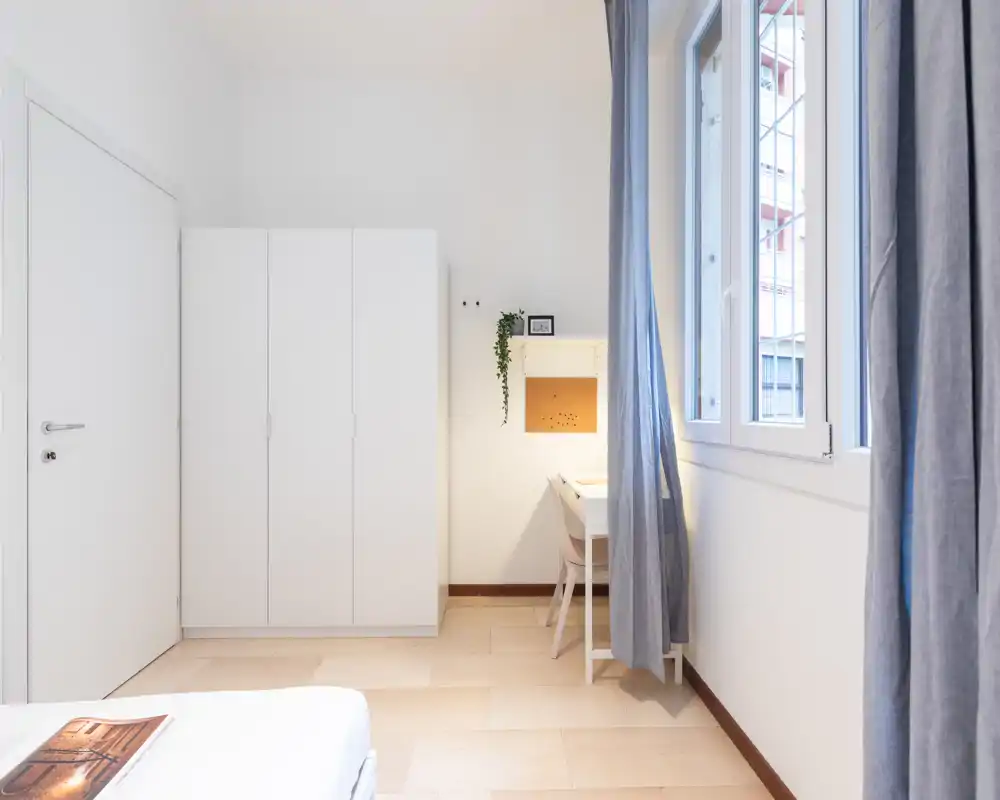 Rincón de dormitorio minimalista y luminoso con la esquina de la cama visible, armario blanco y un pequeño escritorio junto a la ventana que aporta luz natural y estilo moderno.