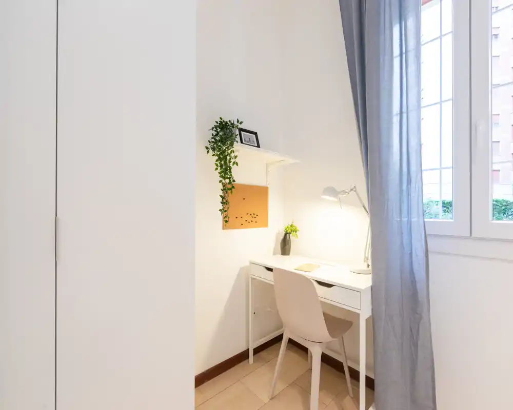 Rincón de dormitorio luminoso con un escritorio blanco compacto y silla junto a la ventana, cortinas grises suaves y un armario a la izquierda — ideal como zona de trabajo en la habitación.
