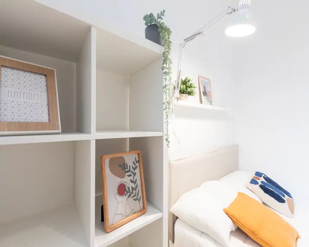 Rincón de dormitorio luminoso y minimalista con cama bien tendida, cojines decorativos y estantería con cuadros y plantas — ideal para una habitación individual o de invitados.