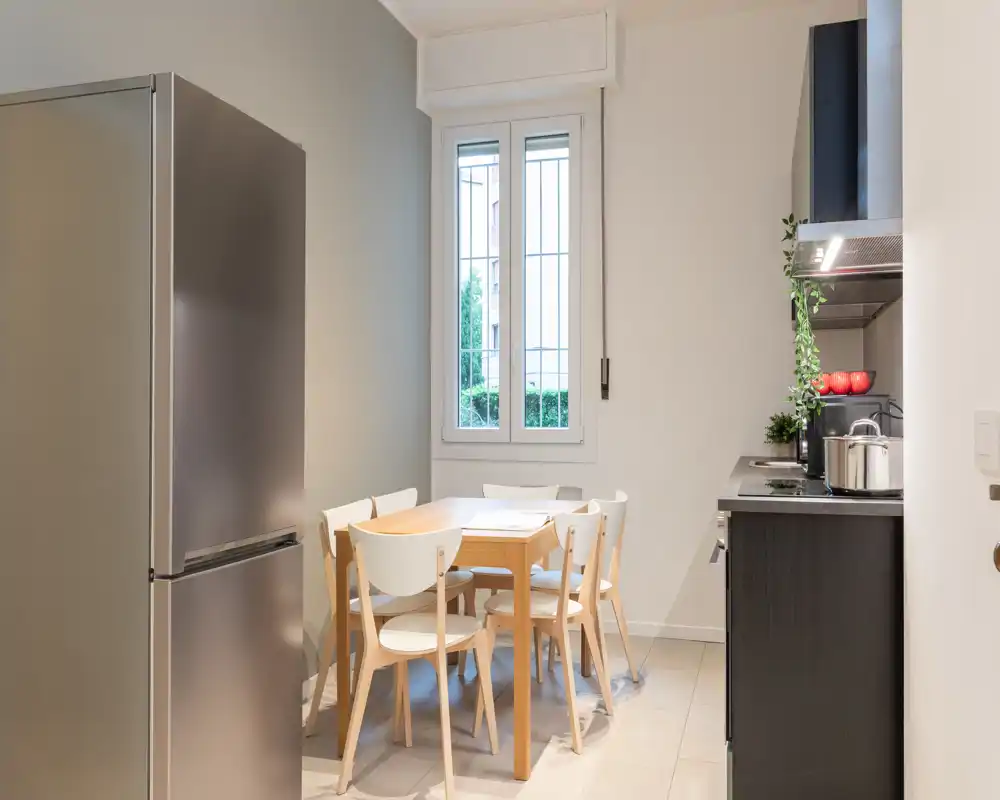 Cucina moderna e compatta con tavolo in legno e quattro sedie, frigorifero in acciaio e mobili scuri; ambiente luminoso e funzionale perfetto per un piccolo appartamento.