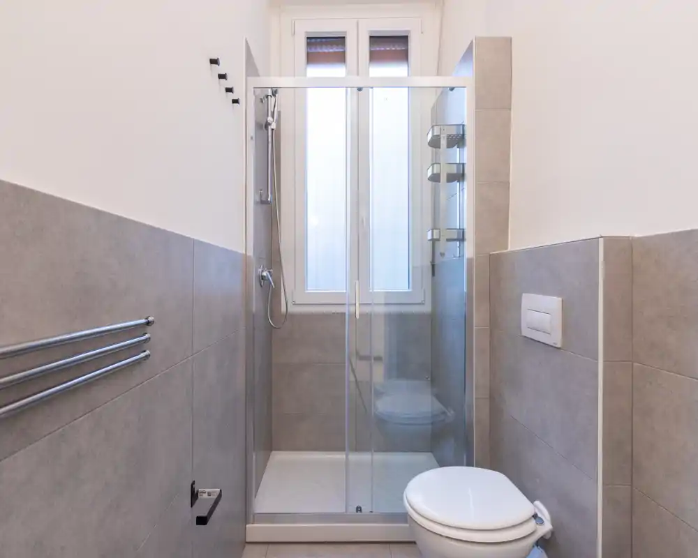 Bagno moderno e pulito con doccia con vetro, wc a muro e piastrelle grigie neutre. La luce naturale dalla finestra valorizza il design minimal.