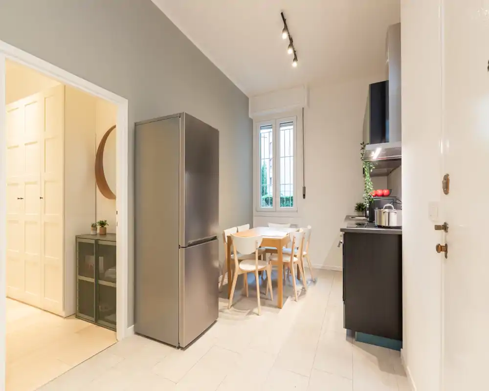 Cucina moderna con frigorifero in acciaio, pensili scuri e un piccolo tavolo da pranzo in legno con sedie; ambiente luminoso, pulito e funzionale.