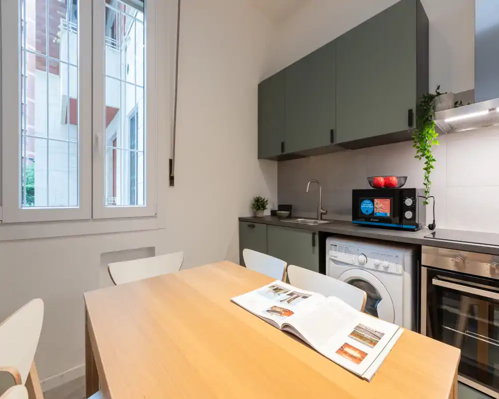 Cucina moderna e luminosa con tavolo in legno, elettrodomestici integrati e lavatrice sotto il piano — styling curato e luce naturale la rendono accogliente.