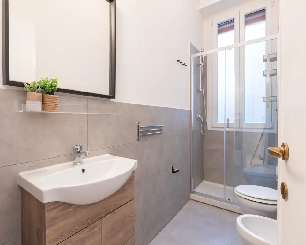 Bagno moderno e luminoso con cabina doccia in vetro, lavabo su mobile e bidet; piastrelle grigie neutre e grande specchio per un aspetto pulito e contemporaneo.