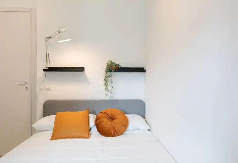 Dormitorio luminoso y minimalista con cabecero gris, ropa blanca y dos cojines ocre; lámpara de pared y estantes flotantes con una pequeña planta le dan un toque acogedor y moderno.