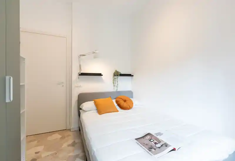 Dormitorio compacto y luminoso con cama doble bien hecha, cojines cálidos y una lámpara de lectura. Estilo moderno y minimalista con paredes blancas y estantes discretos.