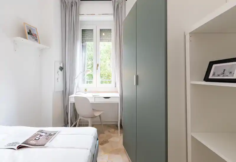 Dormitorio luminoso y compacto con cama blanca, escritorio junto a la ventana y armario alto verde — ideal para una persona o estudiante.