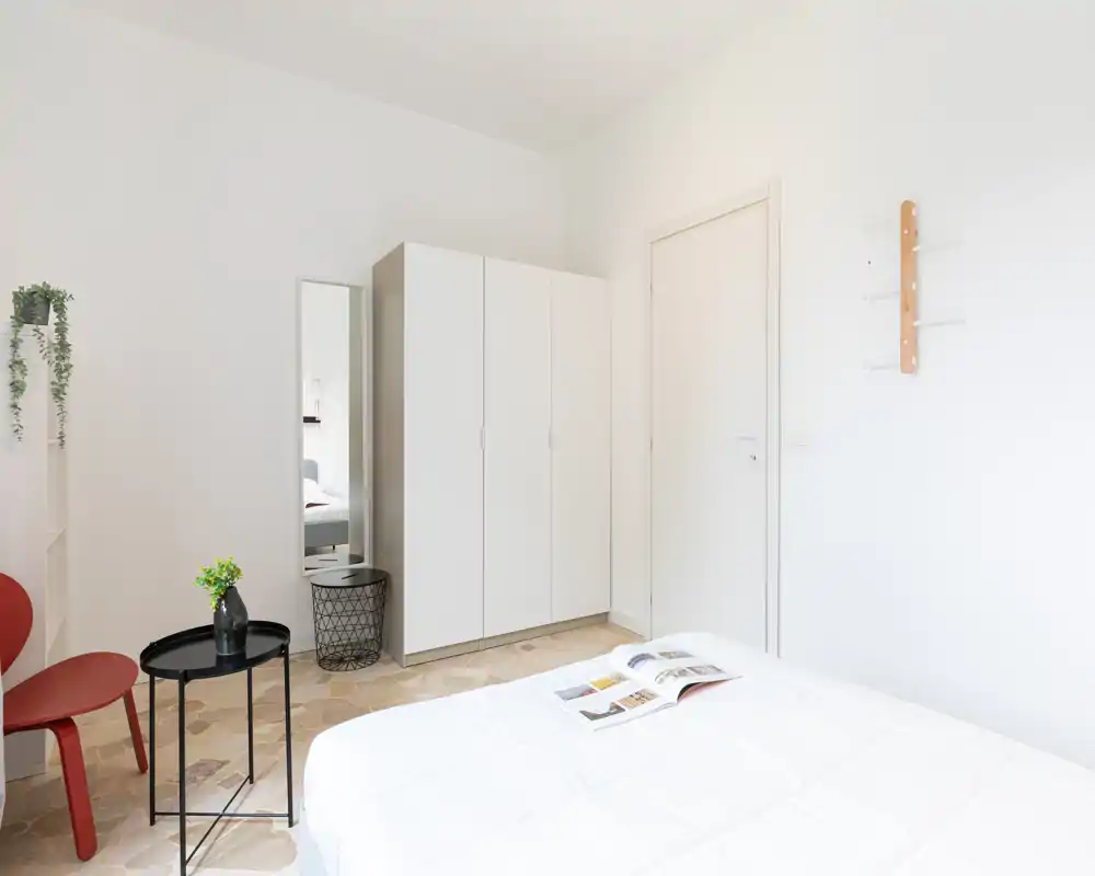 Camera da letto luminosa e minimal con armadio bianco, letto con biancheria bianca e decorazioni semplici come un tavolino nero e una pianta. Linee pulite e luce naturale rendono la stanza ideale per un annuncio moderno.