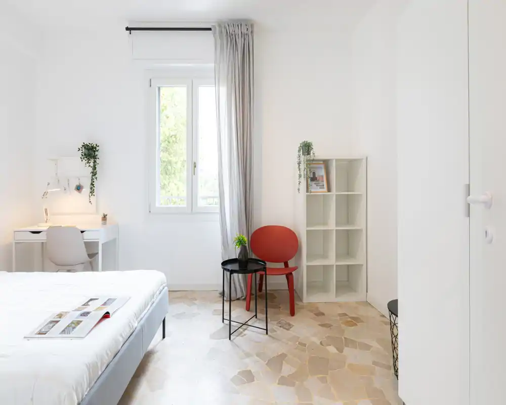 Camera da letto luminosa e minimalista con letto matrimoniale, scrivania vicino alla finestra e un angolo relax con una sedia rossa e tavolini. Toni neutri, luce naturale e decorazioni semplici creano un ambiente tranquillo e accogliente.