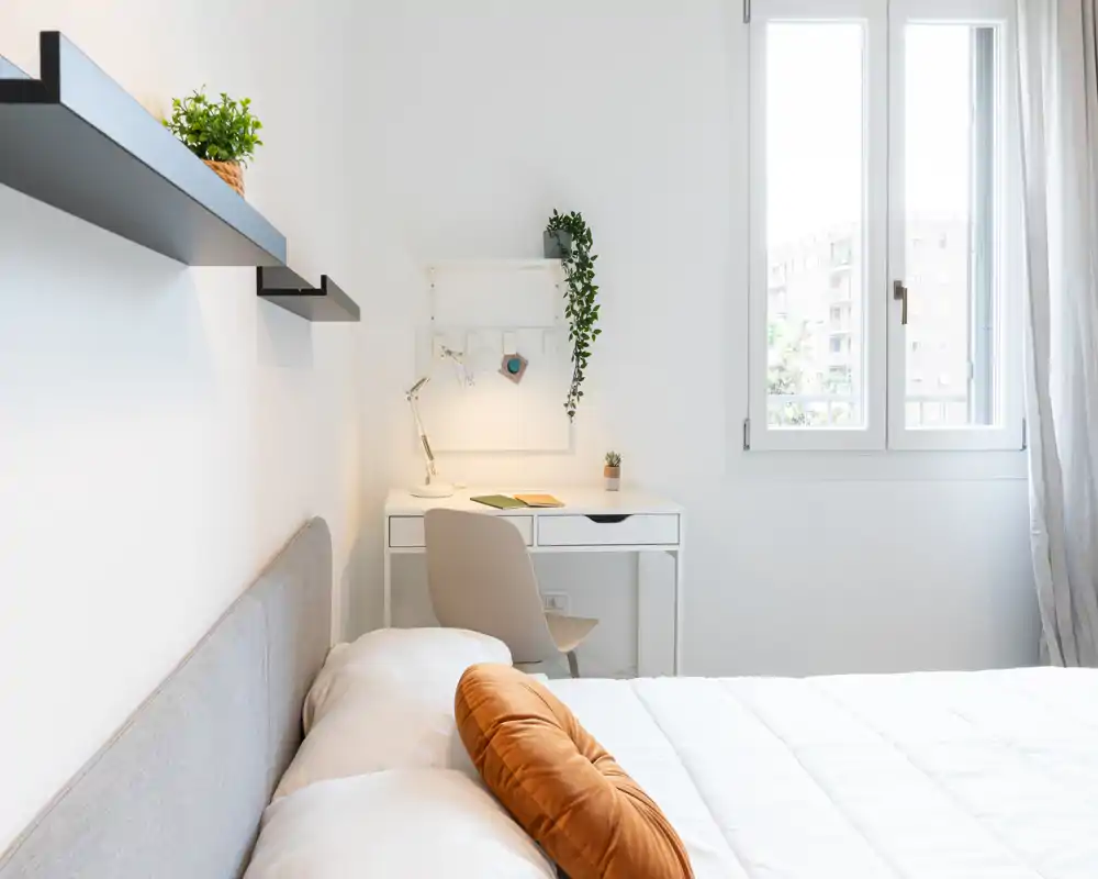 Camera da letto moderna e luminosa con letto comodo, cuscino decorativo rotondo e una piccola scrivania vicino alla finestra, perfetta per riposo e smart working.